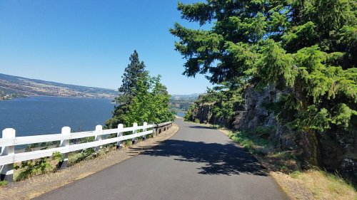 A scenic road lined with trees, overlooking a river and hills under a clear blue sky.