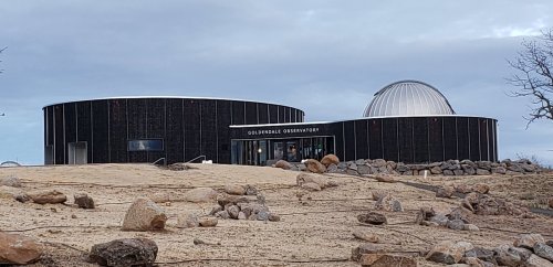 A modern observatory building with a dome, surrounded by rocky terrain and a cloudy sky.