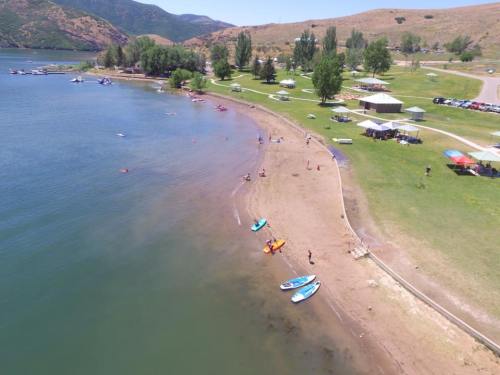 Aerial view of a sandy beach by a lake, with people kayaking, picnicking, and enjoying the sunny day.