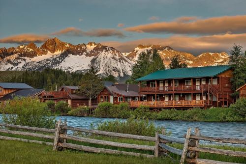 A scenic view of wooden cabins by a river, with snow-capped mountains and a colorful sunset in the background.