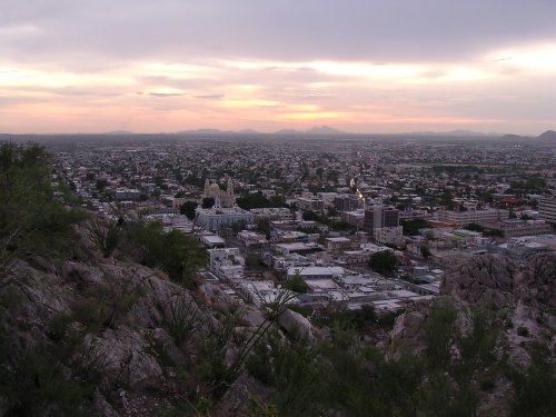 A panoramic view of a city at sunset, with mountains in the background and buildings nestled among greenery.