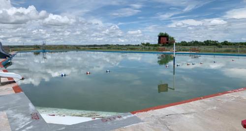 A large, empty swimming pool under a cloudy sky, with reflections on the water and poolside markings visible.