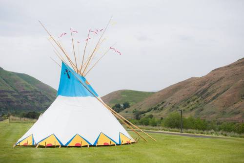 A colorful teepee stands on green grass, surrounded by rolling hills under a cloudy sky.