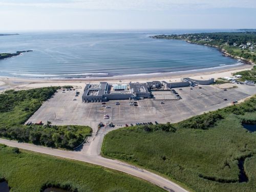Aerial view of a beach with a parking lot and a building near the shore, surrounded by greenery and calm waters.