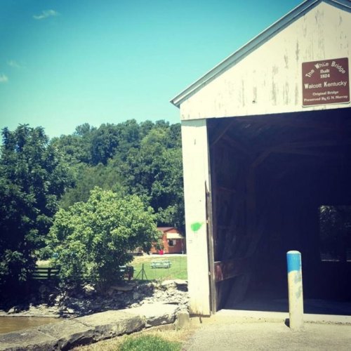 A covered bridge in Walcott, Kentucky, with trees and a river in the background under a clear blue sky.
