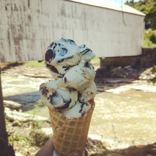 A hand holds a waffle cone topped with scoops of cookies and cream ice cream, with a rustic white building in the background.