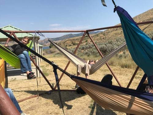 A group of people relaxes in colorful hammocks set up outdoors, surrounded by hills and a calm body of water.