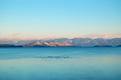 A serene lake scene with calm waters reflecting mountains under a clear blue sky. Snow-capped peaks in the distance.