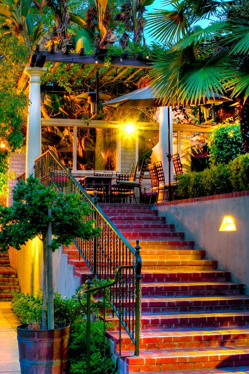 A beautifully lit staircase leads to a patio surrounded by lush greenery and elegant outdoor seating.
