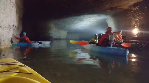 A group of kayakers explores a dimly lit cave, with reflections on the water and rocky walls surrounding them.
