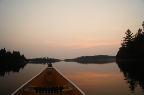A serene lake at sunset, viewed from the front of a canoe, surrounded by trees and soft, colorful skies.