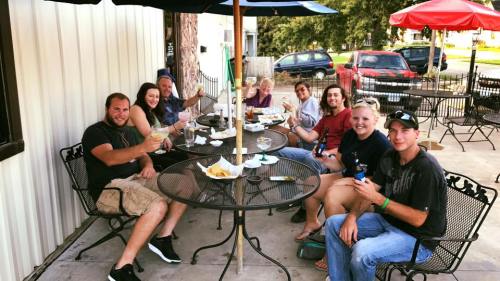 A group of eight people sitting at outdoor tables, enjoying drinks and food, smiling and toasting together.