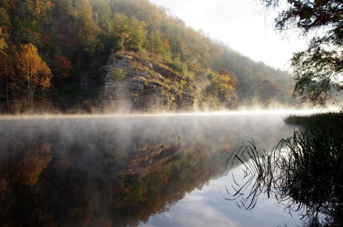 A serene river scene with mist rising, reflecting colorful autumn trees and rocky cliffs in calm water.