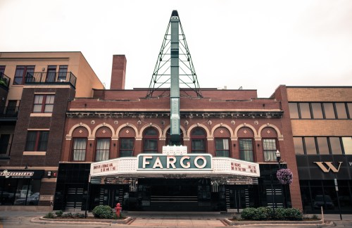 Historic theater building with a marquee displaying "Fargo," surrounded by urban architecture and cloudy skies.