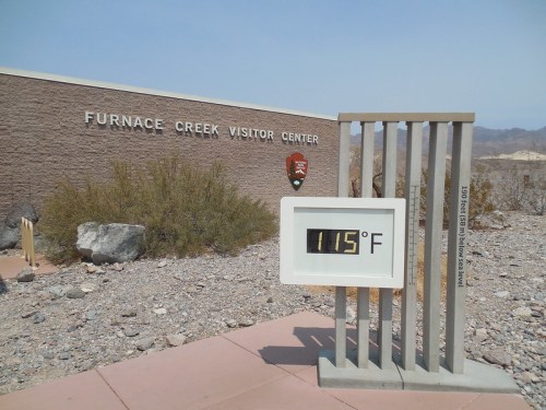 Furnace Creek Visitor Center sign with a temperature display showing 115°F in a desert landscape.