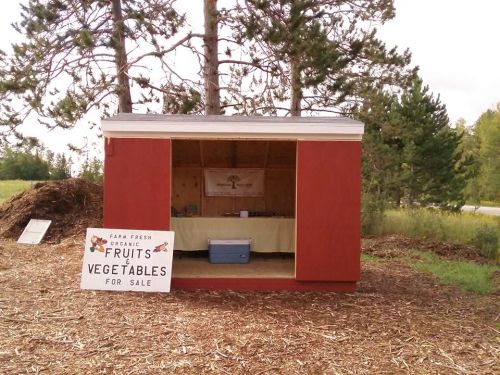 A small shed with a sign reading "Fruits Vegetables for Sale" surrounded by trees and mulch.