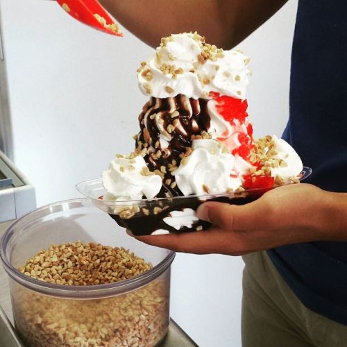 A person holds a large ice cream sundae topped with whipped cream, chocolate syrup, nuts, and a cherry.