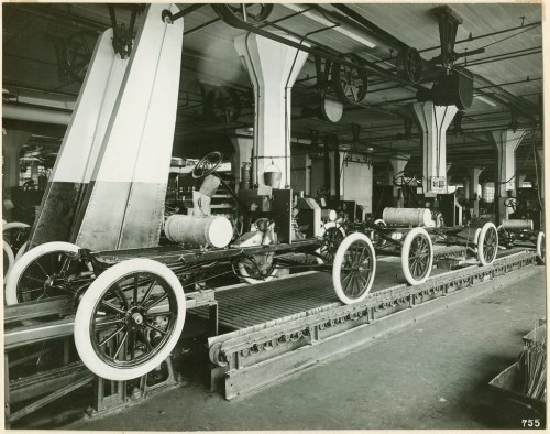 Black and white photo of an early automobile assembly line with car chassis and wheels in a factory setting.
