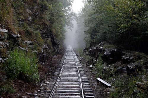 A misty railway track stretches through a dense, green forest, surrounded by rocky terrain and light rain.