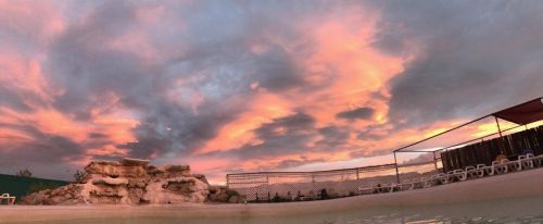 A serene sunset with vibrant pink and orange clouds over a pool area, featuring a rock formation and lounge chairs.