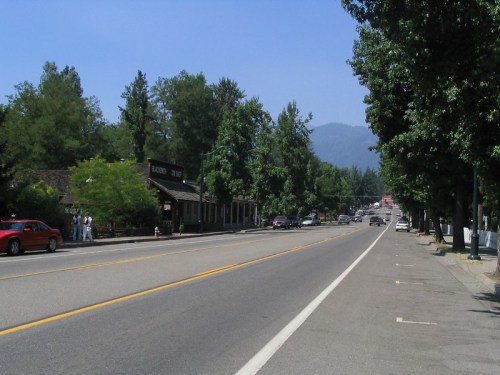 A quiet street lined with trees, featuring a few cars and buildings under a clear blue sky.