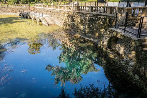 A stone bridge over a clear, reflective pond surrounded by trees and greenery.