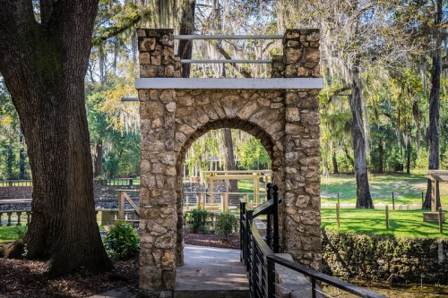 Stone archway with a wooden railing, surrounded by trees and greenery in a serene park setting.