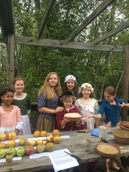 A group of children in historical costumes gather around a table with apples and a pie outdoors.
