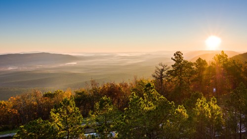 A scenic view of mountains and valleys at sunrise, with trees in the foreground and a golden sky.