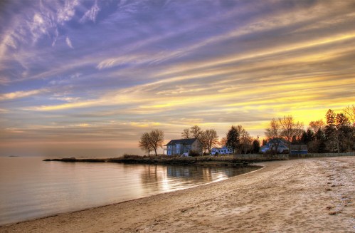 A serene beach at sunset, with calm waters, a sandy shore, and a house surrounded by trees under a colorful sky.