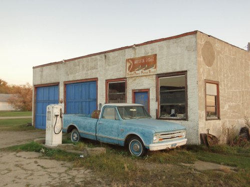 A vintage blue pickup truck parked in front of an old gas station with a weathered building and blue doors.