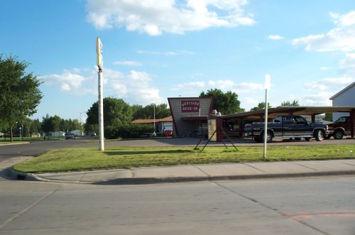 A roadside view of a building with a sign, surrounded by trees and parked cars under a canopy.