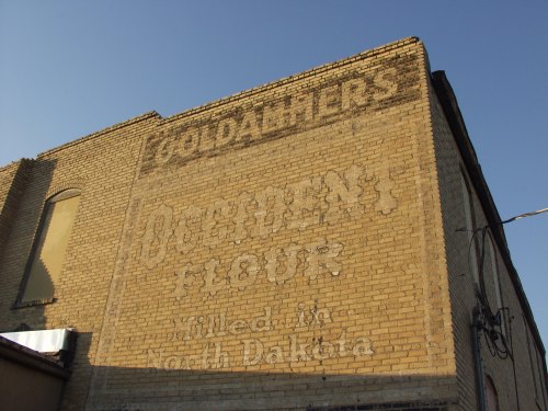 Faded advertisement on a brick wall for "Goldthwait's Flour" from North Dakota, with a clear blue sky above.