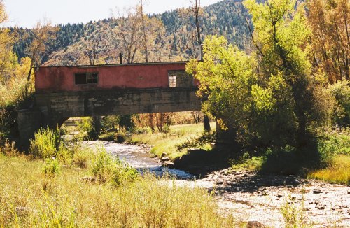 A weathered pink building sits atop a concrete bridge over a flowing stream, surrounded by lush greenery and mountains.