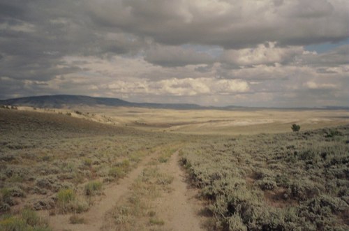 A dirt path winds through a vast, open landscape with rolling hills and a cloudy sky in the background.