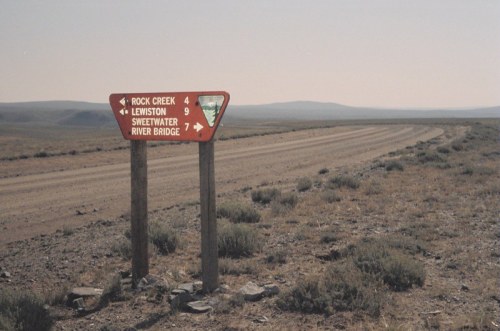 A weathered directional sign on a dirt road, indicating distances to Rock Creek, Lewiston, Sweetwater, and River Bridge.