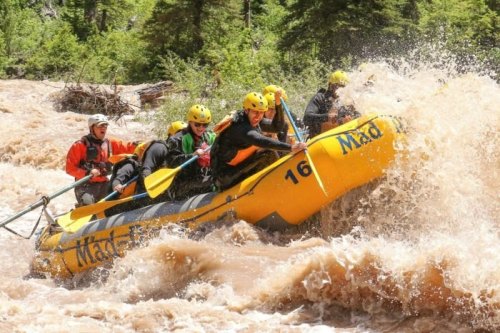A group of people in yellow helmets navigating a white-water raft through turbulent, muddy river rapids.