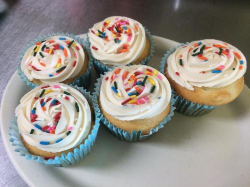 Five frosted cupcakes with colorful sprinkles on a white plate, set against a metallic background.