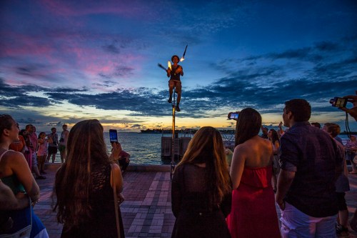 A performer juggles fire on a pole at sunset, while an audience watches and takes photos by the waterfront.