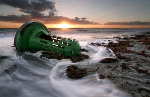 A green buoy lies on a sandy beach, partially submerged in water, with waves gently washing over it at sunset.