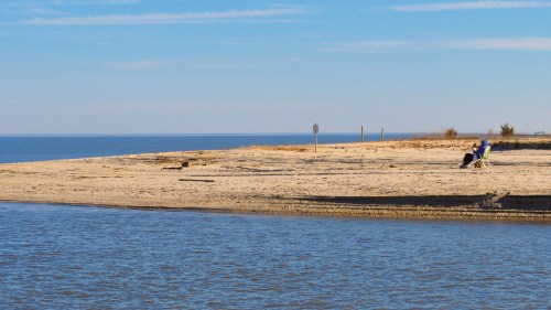 A serene beach scene with two people sitting on chairs by the water, under a clear blue sky.