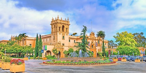 A historic building with ornate architecture, surrounded by lush greenery and colorful flowers, under a cloudy sky.