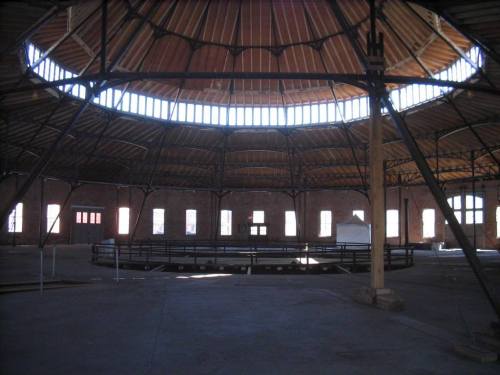 Interior of a large, circular building with a wooden ceiling and open space, featuring a central circular area and brick walls.