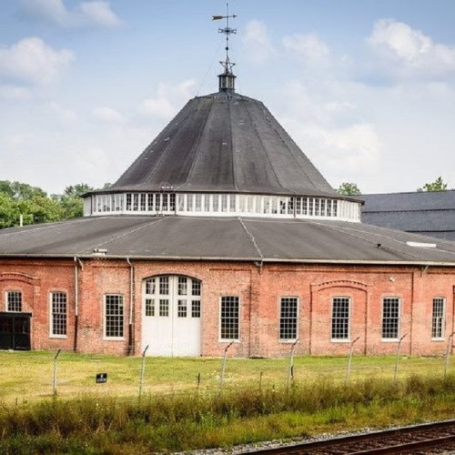 Historic roundhouse with a conical roof, surrounded by grass and a fence, under a blue sky with scattered clouds.