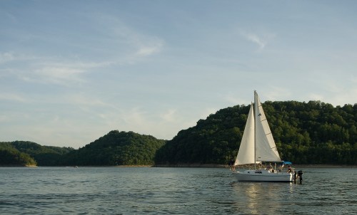 A sailboat glides across calm waters, surrounded by lush green hills under a clear sky.