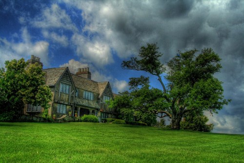 A large, elegant house with multiple windows sits on a green lawn under a dramatic sky with clouds.