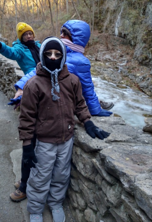A child in winter clothing stands by a stone wall near a flowing stream, with two adults in the background.