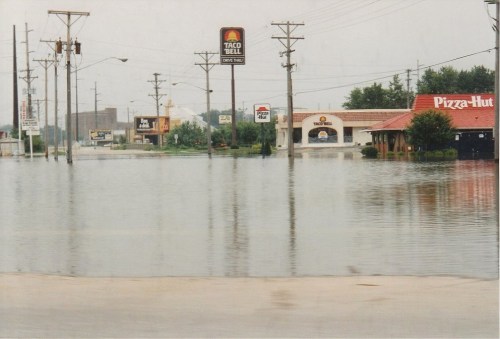 A flooded street with submerged fast-food restaurants and power lines, indicating severe water damage.