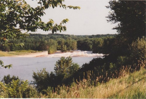 A serene river scene with a dam, surrounded by lush greenery and trees under a clear blue sky.
