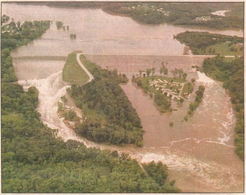 Aerial view of a flooded area with a dam, surrounded by rising water and greenery, showing significant water overflow.